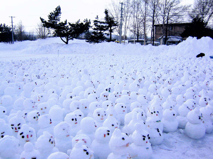 Hundreds of small snowman designs with varied facial expressions densely covering a snowy outdoor area near trees and buildings.