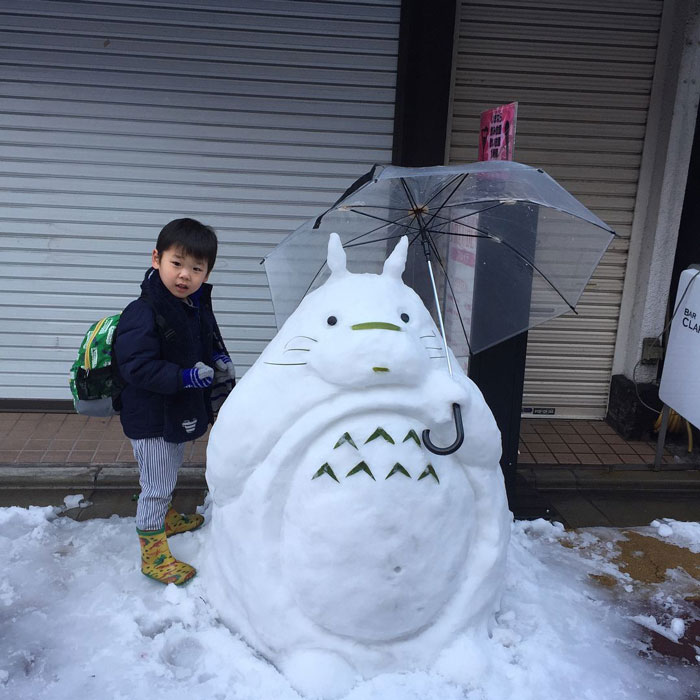 Child standing next to a unique snowman design shaped like a character holding a clear umbrella in the snow.