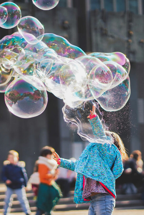 Child in a blue jacket reaching for large soap bubbles outdoors on a sunny day.