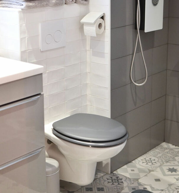 Modern bathroom with a gray toilet seat, white tiles, and patterned floor, humorously cramped.