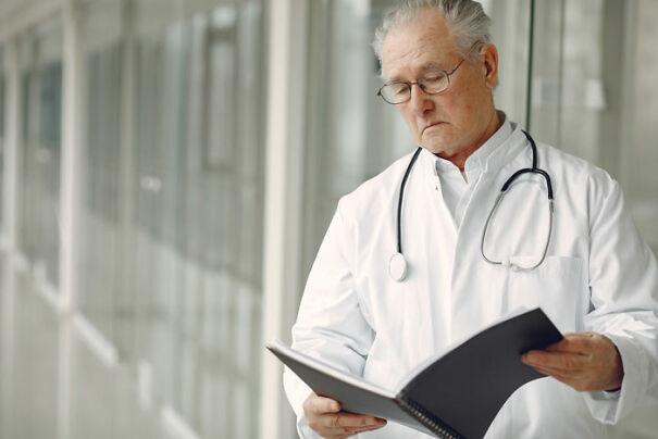 Elderly doctor in a white coat with a stethoscope, reading a notebook in a bright corridor.