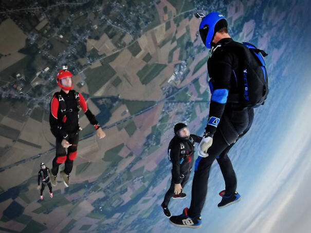 Skydivers in mid-air, wearing colorful suits and helmets, descending above a patchwork of fields and landscape below.