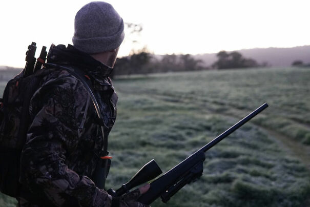 Person in camouflage with a rifle, standing in a frosty field at dawn.