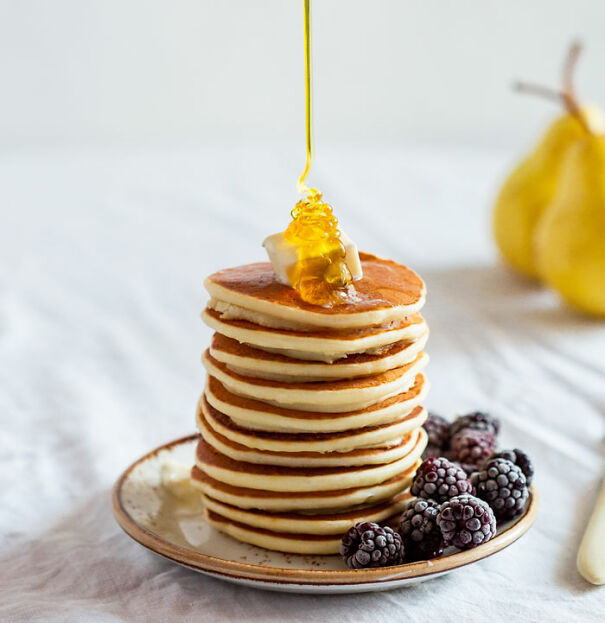 Stack of pancakes with syrup, butter, and blackberries, on a plate with pears in the background, related to dark jokes.