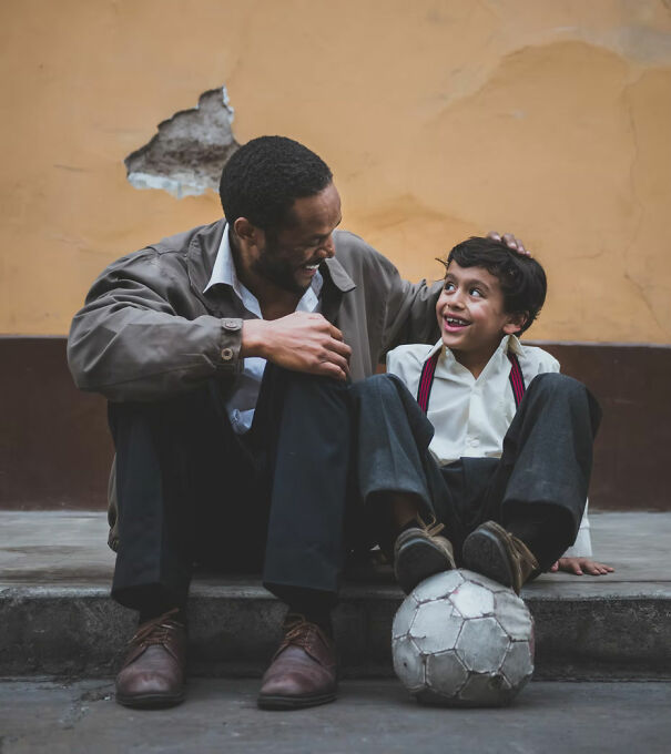 A man and boy sitting on a curb, smiling, with a soccer ball, sharing a light moment.