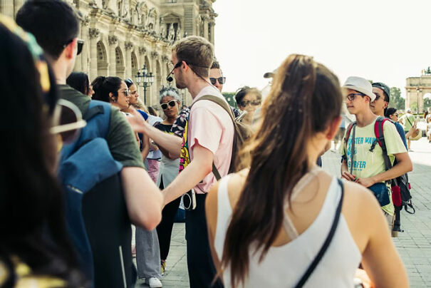 A group of tourists in a sunny plaza, listening to a guide, wearing summer attire and backpacks.