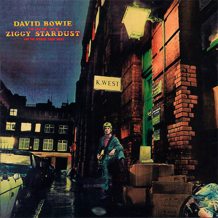 Greatest album cover featuring a figure in a vibrant outfit in a dimly lit urban street with a brick building.