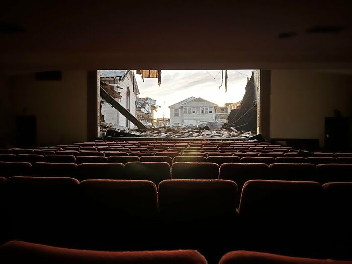 Empty theater seats facing a large hole showing Mayfield tornado aftermath with damaged buildings and debris outside. Empty theater seats facing a large hole showing Mayfield tornado aftermath with damaged buildings and debris outside.