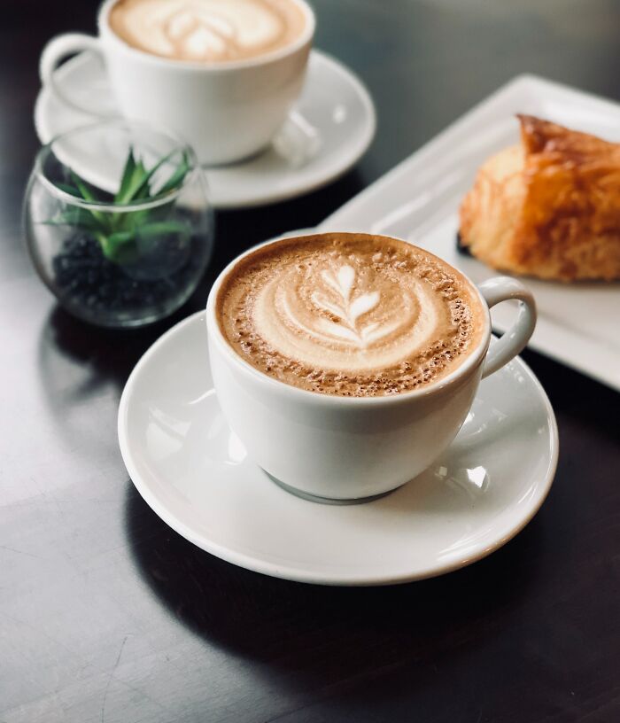 Two cups of latte with latte art, a small plant, and a croissant on a table related to weird and disturbing dates.