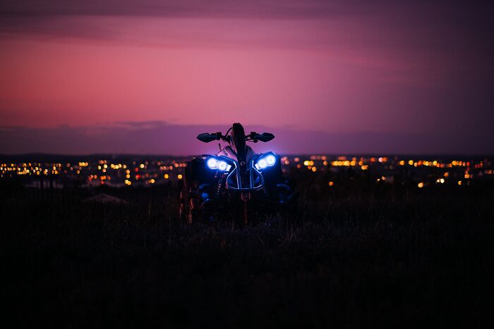 All-terrain vehicle with bright headlights on a dark field at dusk, city lights blurred in the background.