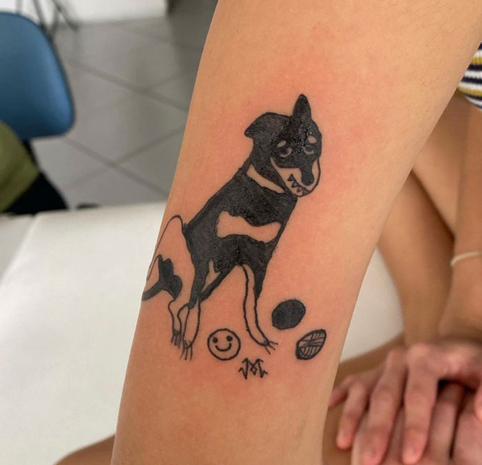 Tattoo of a cartoon-style dog and symbols on arm by artist known for unique drawing style.