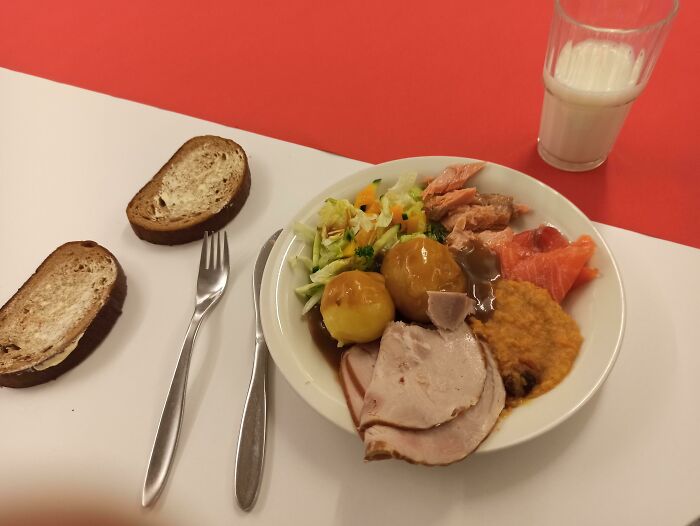 School lunch featuring sliced meat, potatoes with gravy, mixed salad, bread slices, and a glass of milk on a red and white table.