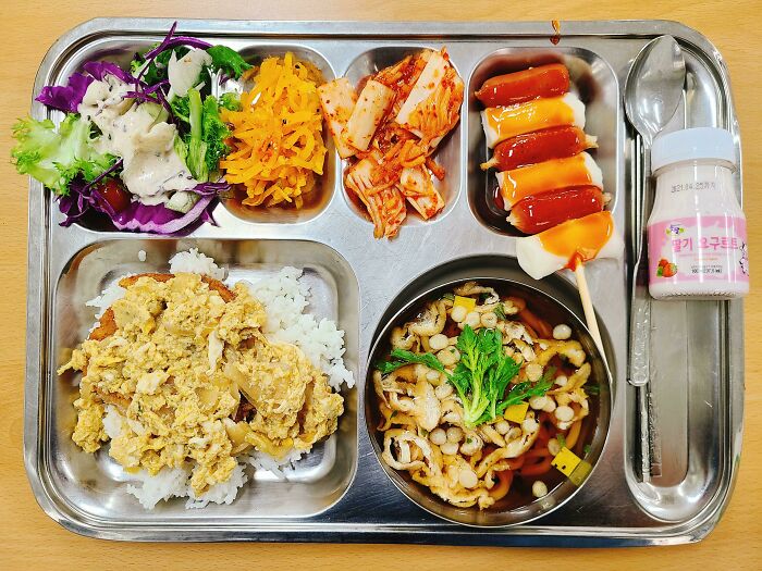 Korean school lunch tray with rice, egg dish, kimchi, salad, soup, skewered sausages with sauce, and a drink yogurt.