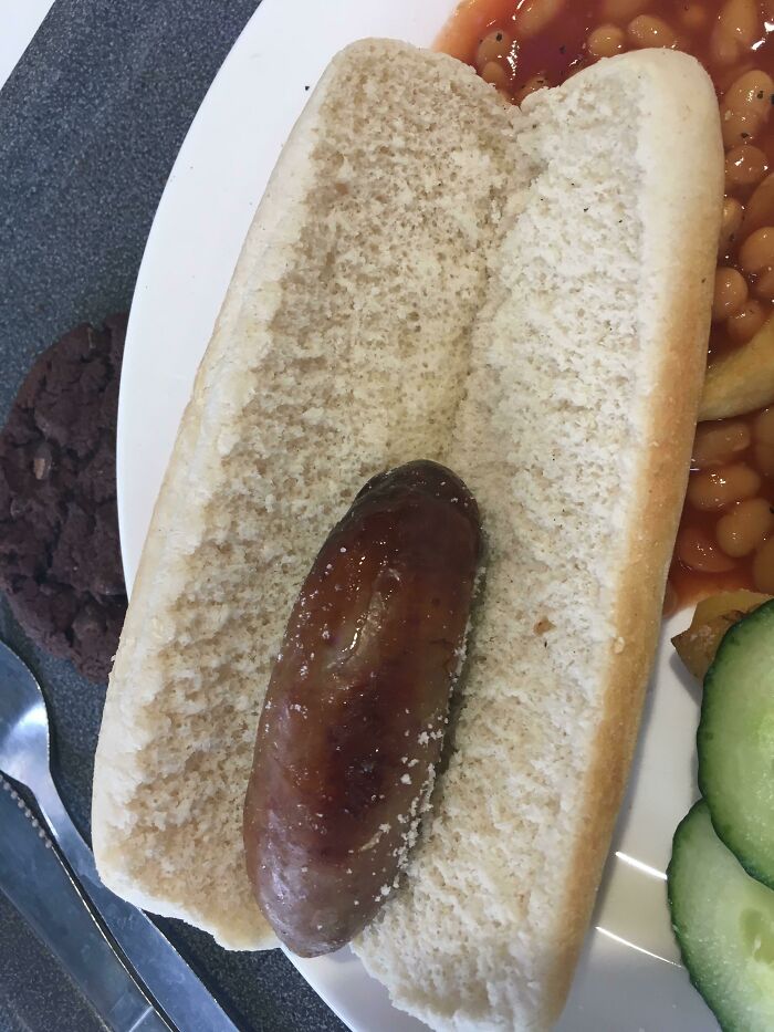 Simple school lunch with sausage in a bun served alongside baked beans, cucumber slices, and a chocolate cookie on a white plate.