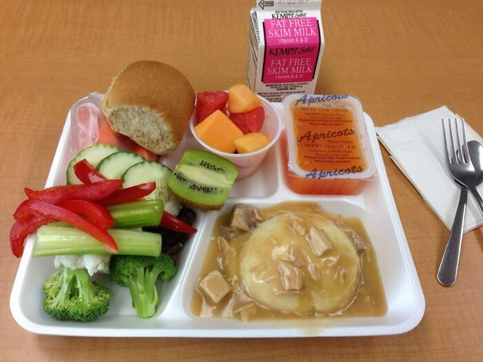 School lunch tray with mashed potatoes and gravy, fresh vegetables, fruit cup, roll, and fat-free skim milk.