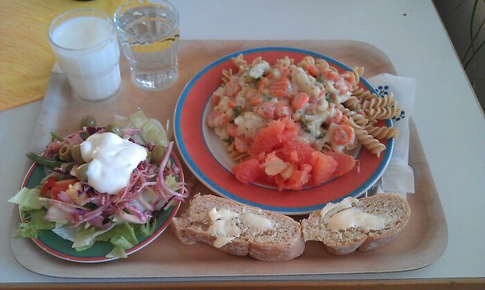 School lunch tray with pasta, salad topped with dressing, tomato slices, buttered bread, milk, and water glass from around the world.