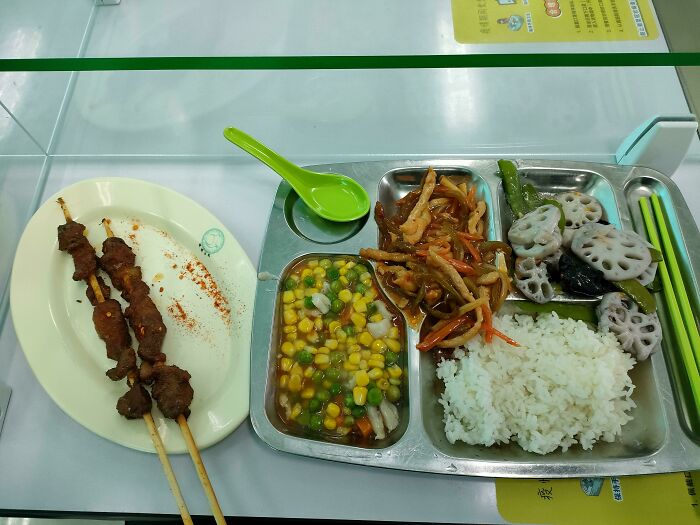 School lunch featuring skewered meat, mixed vegetables, rice, and lotus root served on a divided metal tray.