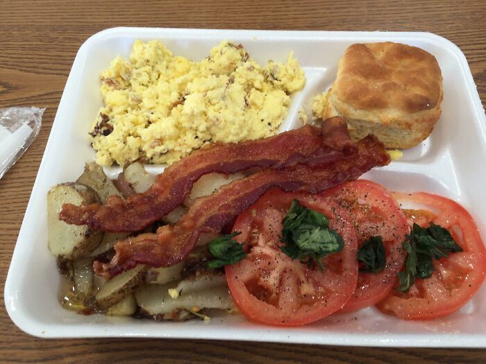 American school lunch plate with scrambled eggs, bacon, potatoes, sliced tomatoes, and a biscuit on a wooden table.
