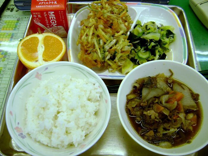 Japanese school lunch tray with rice, vegetable tempura, seaweed salad, stew, orange halves, and a carton of milk.