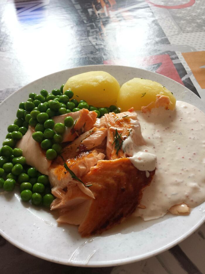 School lunch with salmon, boiled potatoes, green peas, and creamy sauce on a white plate on a table.