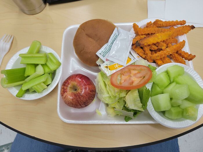 School lunch featuring a sandwich, sweet potato fries, celery, apple, lettuce with tomato, and melon slices on a tray.
