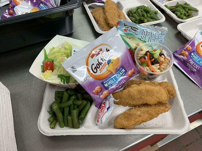 School lunch tray with chicken tenders, green beans, salad, pasta veggie cup, apple slices, and a snack bag of pretzels.