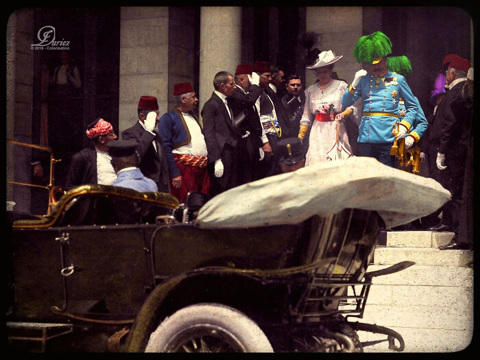 Historical figures in vibrant attire exit a building with a vintage car in the foreground.