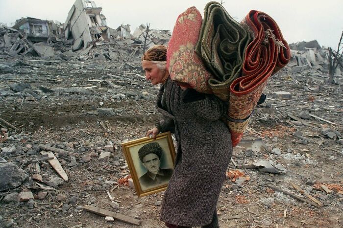Woman carrying rugs and a portrait in a war-torn area, depicting an important historical moment.