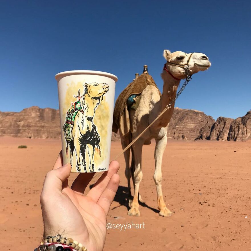Hand holding paper cup with camel illustration against a real camel in desert, showcasing travel-themed cup art.