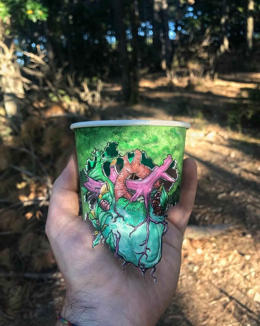 Hand holding a paper cup with a detailed colorful illustration of an anatomical heart outdoors in a forest setting.