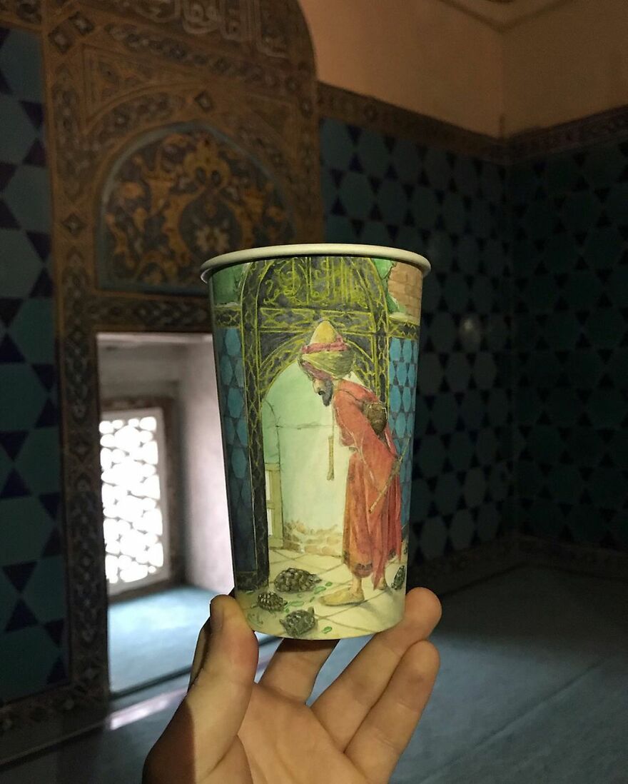 Hand holding a paper cup with detailed travel-inspired illustration of a person and turtles inside an ornate room.