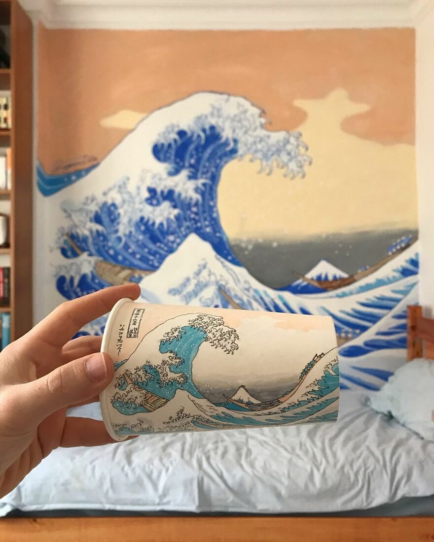 Hand holding a paper cup with detailed wave illustration matching the large wall art behind, showcasing artist's travel drawings.