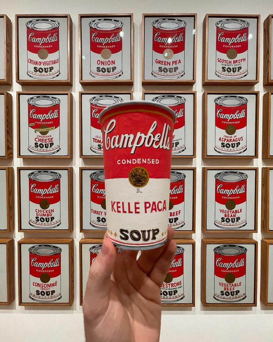 Hand holding a paper cup with Campbell’s soup illustration, displayed in front of framed soup can art on a wall.