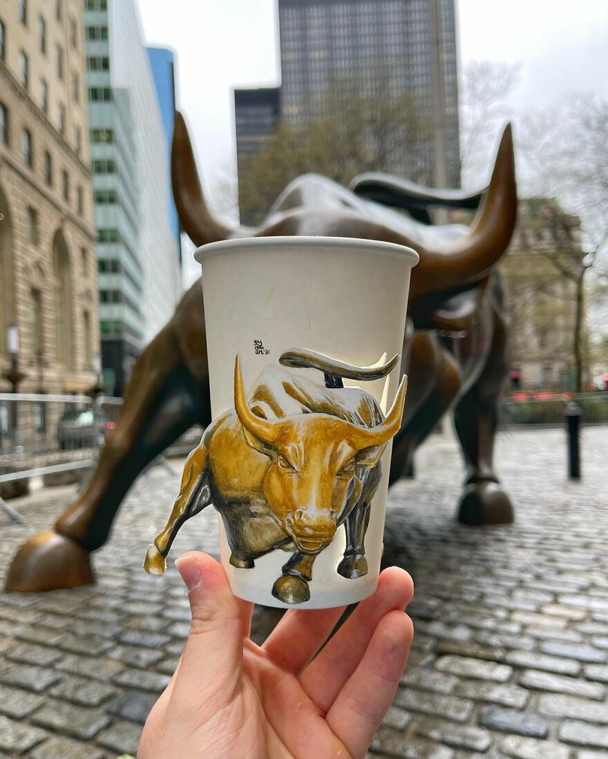 Paper cup illustration of a charging bull held in front of the actual bull statue on a city street during travel.