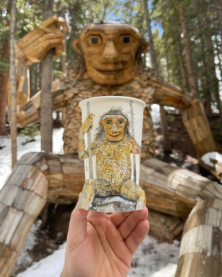 Hand holding paper cup with detailed illustrated monkey matching large wooden monkey sculpture in forest.