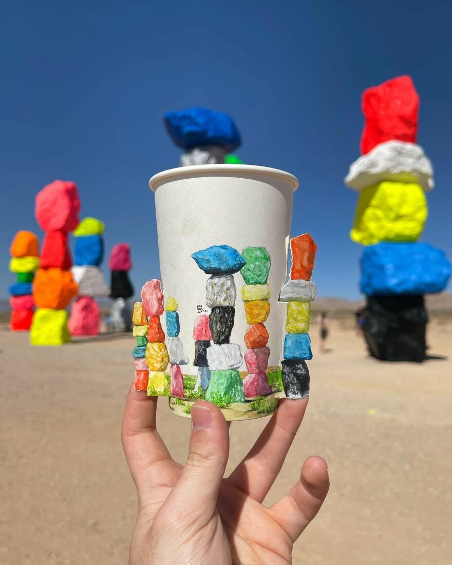 Hand holding a paper cup with colorful stacked rock illustrations drawn while traveling the world in a desert setting.