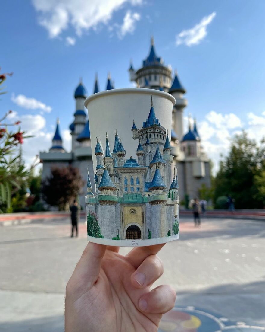 Hand holding a paper cup with detailed castle illustrations matching the real castle in the background, showcasing travel art.