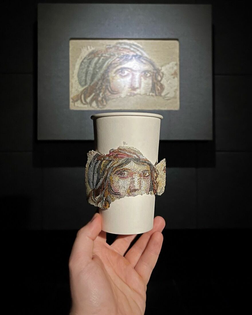 Hand holding a paper cup with detailed illustration of a face drawn during traveling, museum artwork in background.