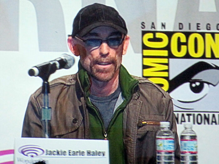 Actor at Comic-Con panel discussing career revival roles, wearing sunglasses and a cap behind a microphone.