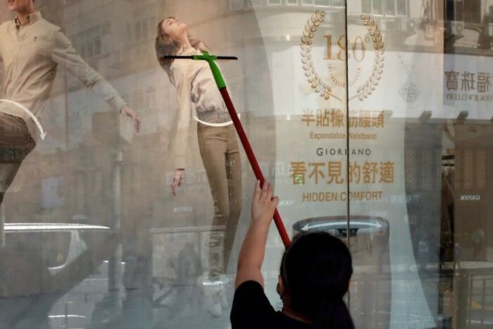 Person cleaning a glass window with a reflection showing a woman leaning back, a lucky accident captured by photographer Edas Wong.