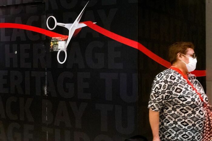 A person wearing a mask walking past a mural showing large scissors cutting a red ribbon, a lucky accident captured by Edas Wong.