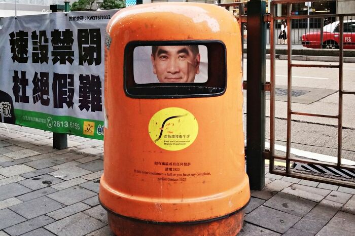Face perfectly aligned with trash can window, capturing a lucky accident by photographer Edas Wong on a city sidewalk.