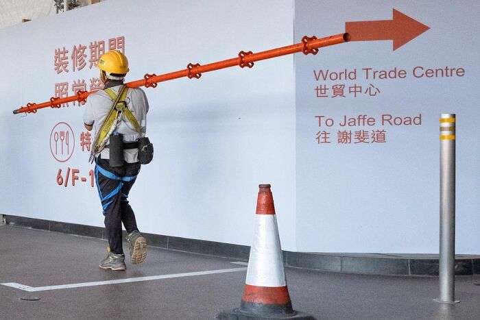 Construction worker carrying a long orange pipe perfectly aligned with a directional arrow in a lucky accident photo.