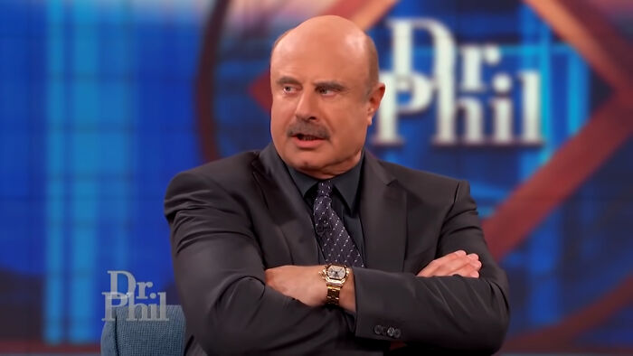 Dr. Phil sitting with arms crossed on set, discussing reality TV and how fake it all was in a studio setting.