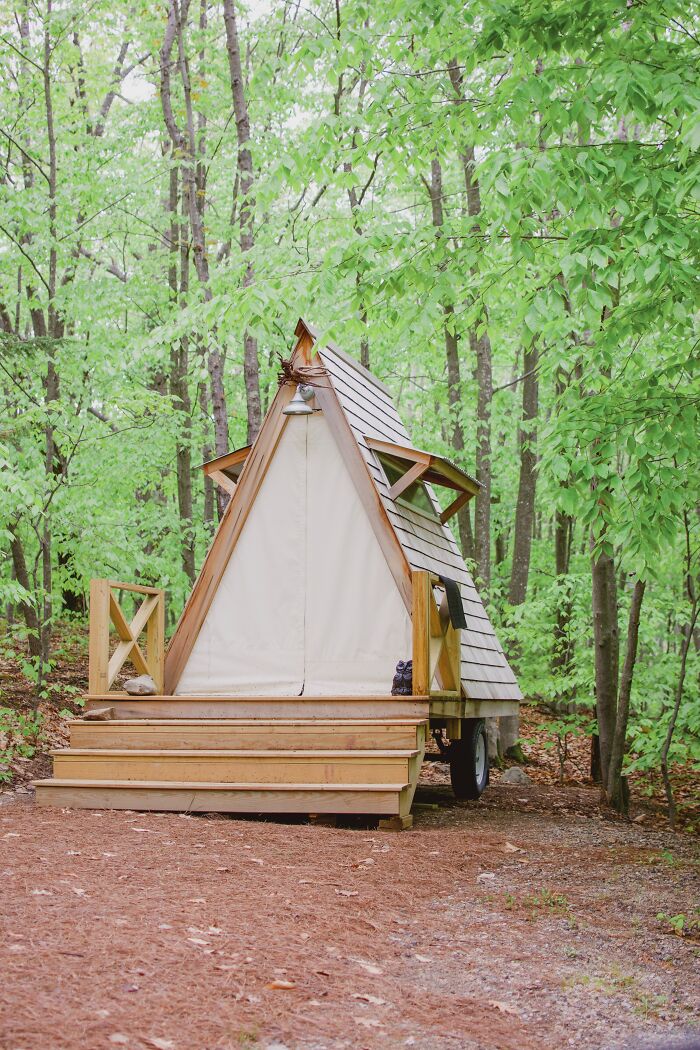 A small wooden A-frame cabin in a forest setting, surrounded by green trees and natural foliage.