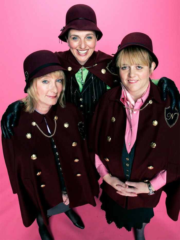 Three women dressed in matching vintage outfits, smiling and posing against a pink background, reality TV cast.