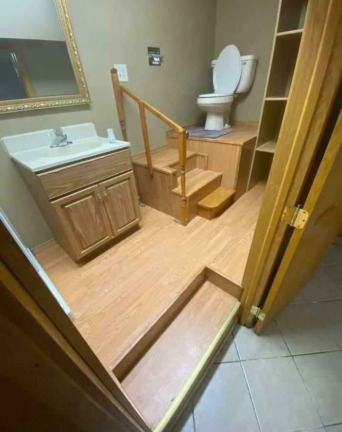 Awful home interior with a toilet on a raised platform, accessible by small wooden stairs next to a sink.