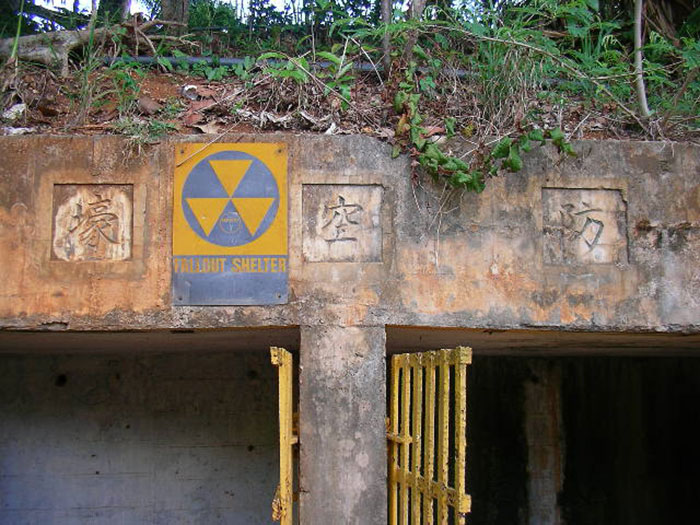 A weathered fallout shelter entrance with a yellow warning sign and overgrown vegetation above, linked to real conspiracies.