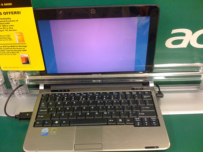 Laptop displaying a blank blue screen, placed on a store counter with promo sign and visible Acer branding.