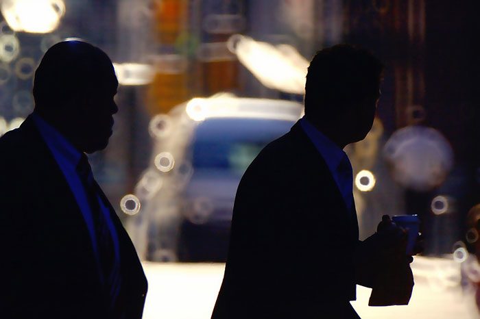 Silhouettes of two men in suits walking with blurred city lights, evoking the theme of conspiracies proven real.